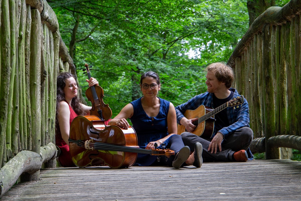 3 Personen sitzen mit Musikinstrumenten auf einer Holzbrücke im Wald